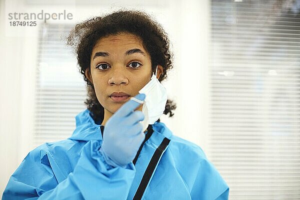Porträt eines jungen  müden afroamerikanischen medizinischen Mitarbeiters oder einer Krankenschwester in Schutzkleidung  die ihre Gesichtsmaske abnimmt und mit frustriertem Gesichtsausdruck in die Kamera schaut. Mitarbeiter im Gesundheitswesen während der COVID 19 Pandemie