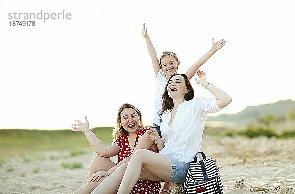 Glückliche Familie mit zwei jungen Frauen und einem kleinen Mädchen  die fröhlich ihre Zeit draußen in der Natur verbringen  am Meeresufer sitzen  lachen und vor Glück schreien  die Hände hochheben. Sommerurlaub