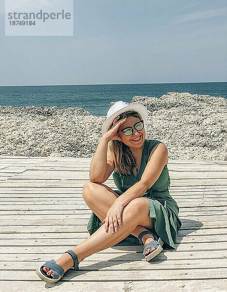 Happy freudige Frau trägt Sonnenbrille und Hut genießen Sommerurlaub Reise am Meer  lächelnd weiblichen sitzen auf hölzernen Terrasse am Strand am sonnigen Tag mit Seelandschaft auf Hintergrund