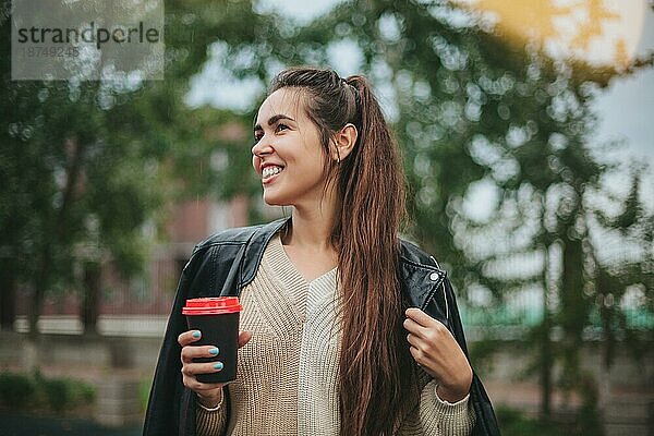 Junge Frau mit Kaffee zum Mitnehmen in der Hand in lässiger Kleidung am Flusswasser am Herbsttag mit Stadtlandschaft im Hintergrund  romantisch träumende Frau an der Küste  die Erholung in der Natur genießt