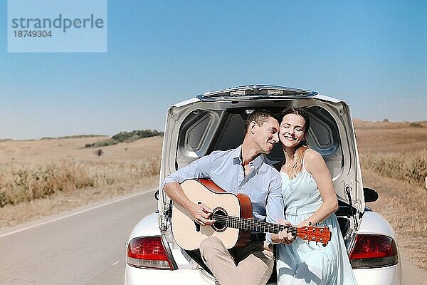 Glückliches junges Paar spielt Gitarre und genießt die gemeinsame Zeit  während es auf dem offenen Kofferraum eines auf einer Landstraße geparkten Autos sitzt