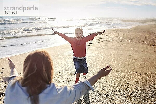 Junge liebende Mutter streckte die Arme zur Seite der lächelnden Tochter aus  die ihr am sonnigen Strand entgegenlief. Die glückliche Mutter ist bereit  das kleine Mädchen zu umarmen und genießt die gemeinsame Zeit am Sommerstrand. Rückansicht