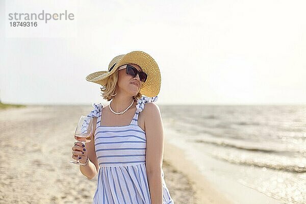 Fröhliche adultes Weibchen in Sommerkleidung lächelt und schaut weg  während sie am Sandstrand in der Nähe des Meeres Wein genießt