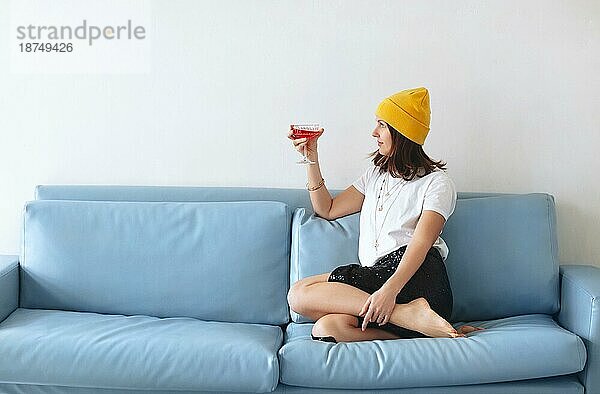 Abgeschnittene Aufnahme einer jungen lächelnden Frau  die ein Cocktailglas mit rotem Alkohol in der Hand hält und zu Hause auf dem Sofa sitzt. Sie trägt modische Kleidung und Accessoires und genießt eine Party mit Freunden im Haus
