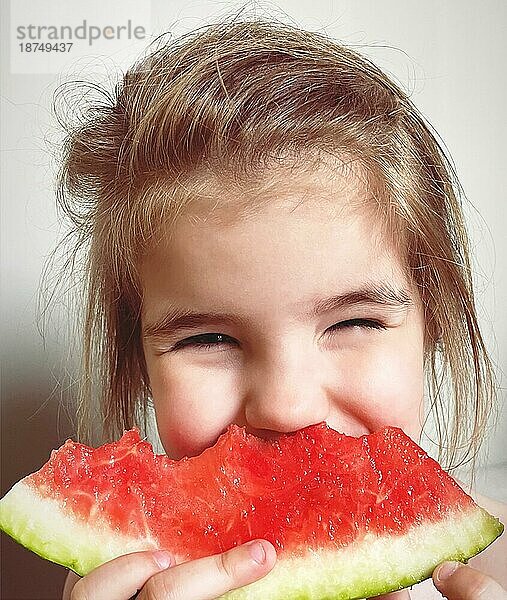 Close up Porträt von glücklichen niedlichen kleinen Mädchen Kind essen geschnitten Scheibe süße Wassermelone und lächelnd in die Kamera mit verengten Augen