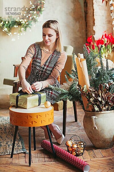 Lächelnde glückliche blonde Frau sitzt auf einem Stuhl im neuen Jahr festlich dekorierten Wohnzimmer und Verpackung Weihnachtsgeschenkbox mit Geschenkpapier und Band Dekor. Konzept der Vorbereitung für die Feiertage