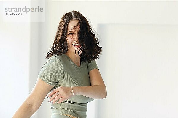 Foto in Bewegung von fröhlicher junger Frau  die springt oder Sport in der Halle treibt  mit breitem Lächeln in die Kamera schaut  während sie sich bewegt  und vor einem unscharfen Wohnungshintergrund posiert