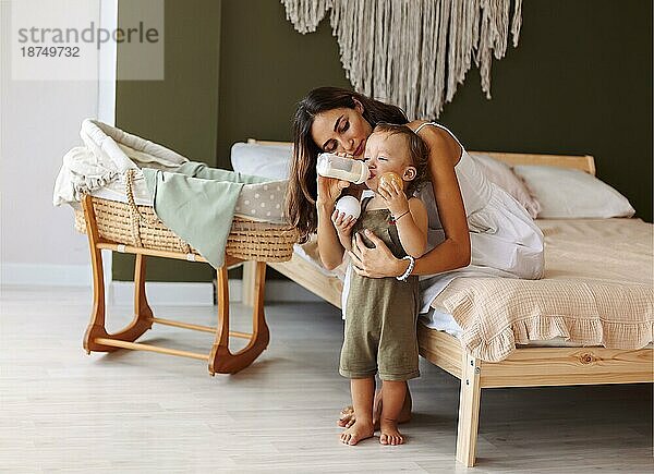 Junge liebevolle Mutter hält Kind Hände und fressen Baby Junge Sohn mit Milch aus der Flasche  schöne Mutter in gemütlichen Boho Schlafzimmer zu Hause und genießen Mutterschaft