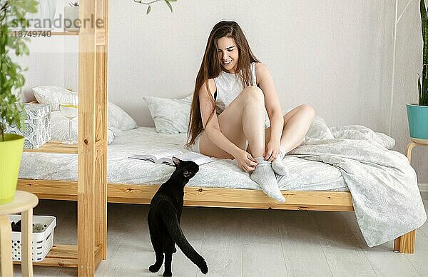 Junge Frau in Ganzkörper Schlafanzug lächelt und spielt mit schwarzer Katze auf dem Bett sitzend am Wochenende morgens zu Hause