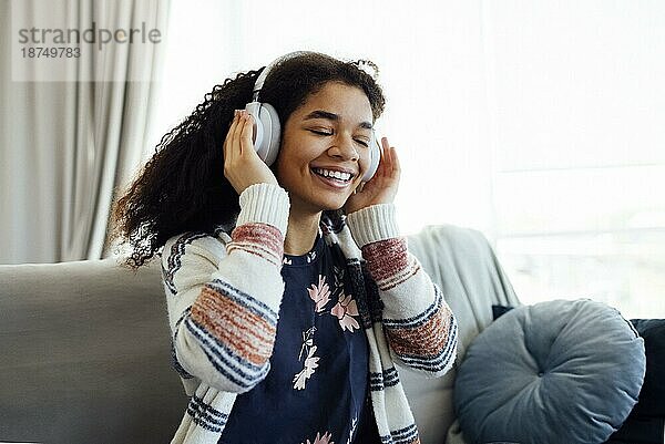 Afrikanische Millennial Frau im orangefarbenen Pullover  die weiße drahtlose Kopfhörer trägt  Musik hört  sich ausruht und auf der Couch im Wohnzimmer liegt. Mädchen verbringen faul Wochenende zu Hause