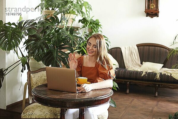 Fröhliche Frau  die an einem klassischen Tisch in einem hellen Wohnzimmer sitzt und mit der Hand am Laptop winkt  während sie online telefoniert