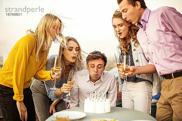 Junger glücklicher Mann bläst Kerzen aus und wünscht sich auf dem Dach einen Geburtstag  umgeben von fröhlichen Freunden
