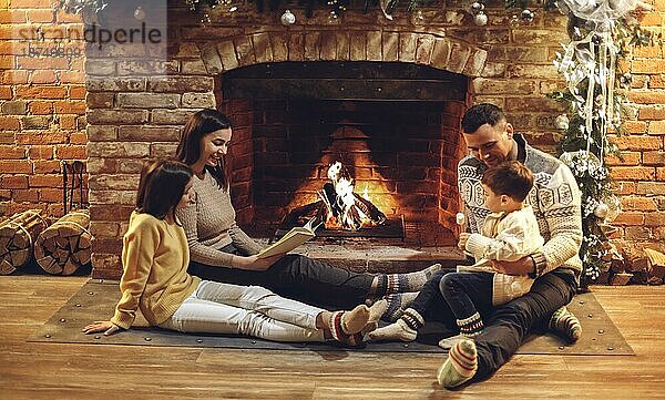 Glückliche europäische Familie mit Kindern  die auf dem Boden vor dem Kamin sitzen und lesen und Marshmallows über dem Feuer braten  junge liebende Eltern mit zwei kleinen Kindern  die den Winterurlaub in einem gemütlichen Landhaus genießen