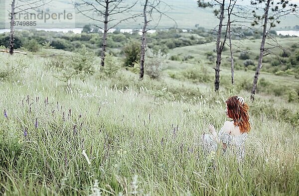 Seitenansicht einer jungen Frau  die ihr rothaariges Haar berührt und sich ausruht  während sie im Gras an einem Sommertag in der Natur sitzt