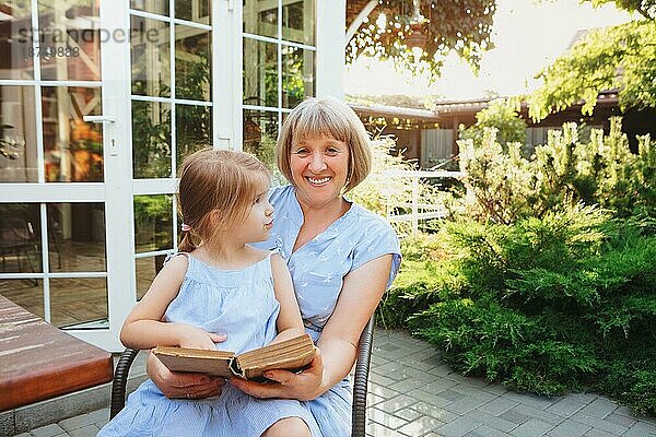 Ältere Großmutter und Enkelin verbringen Zeit zusammen auf der sonnigen Terrasse in der Nähe von Haus  Oma liest Märchen laut zu reizenden Mädchen  während kleines Mädchen sitzt auf ihrem Schoß Sommerurlaub mit Großeltern
