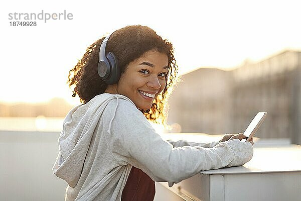 Seitenansicht Porträt der glücklichen fröhliche afrikanische amerikanische Frau in Kopfhörer hören Musik auf dem Smartphone und lächelnd in die Kamera während der Morgengymnastik auf dem Dach