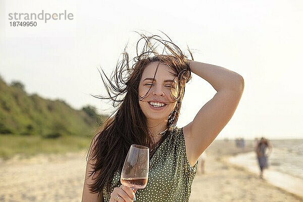 Glückliche adultes Weibchen in Sommerkleidung lächelt und schaut in die Kamera  während sie am Sandstrand in der Nähe des Meeres Wein genießt