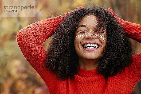Junge glückliche überglückliche afroamerikanische Frau mit lockigem Haar  die Augen geschlossen haltend  lachend und Spaß habend  während sie im Herbstwald steht und die Freizeit in der herbstlichen Natur genießt