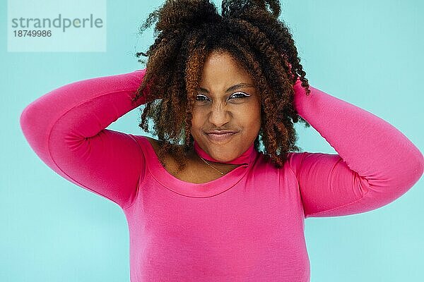 Verträumte junge schöne afroamerikanische Frau mit hellen Augenlinien  die einen rosafarbenen Body über einer blaün Wand trägt  hält die Hände unter dem Kinn zusammen  schaut mit glücklichem Gesichtsausdruck  hat ein zahniges Lächeln
