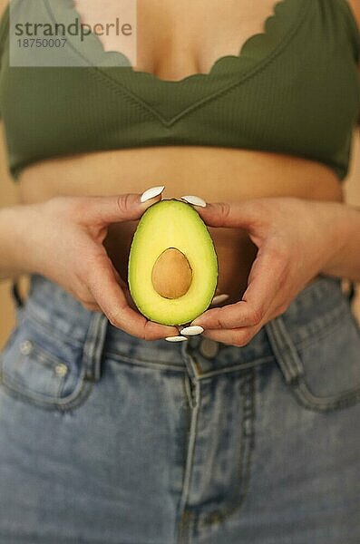 Junge Frau  die in die Kamera schaut und die Hälften einer reifen Avocado vor einem beigen Hintergrund zeigt