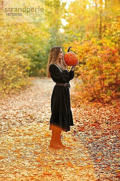 Junge blonde Frau hält gruselig gebastelten Kürbis vor ihrem Gesicht  stehend isoliert über Herbst Wald Hintergrund. Herbstferien  halloween Dekorationen und Menschen Konzept