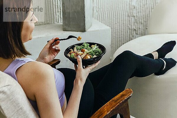 Schwangere junge brünette Frau ißt gesunden Salat aus Schüssel Draufsicht. Gesundes Essen beim Mittagessen auf einem gemütlichen Sessel