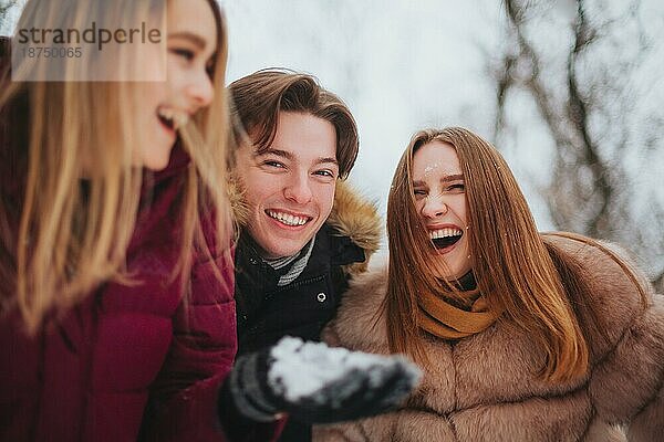 Gruppe von vier fröhlichen besten Freunden  die das kalte Wetter und den ersten Schnee genießen  während sie im Freien über schneebedeckten Bäumen stehen  die Hände hochheben und versuchen  fallende Schneeflocken aufzufangen  und mit einem glücklichen Lächeln nach oben schauen