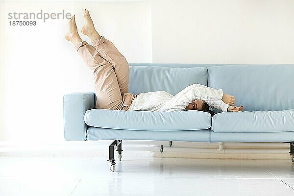 Foto einer jungen  fröhlichen Frau in Freizeitkleidung  die sich mit hochgelegten Beinen auf ein blaues  gemütliches Sofa fallen lässt und spielerisch unter ihrem Arm in die Kamera blickt  um die freie Zeit zu Hause zu genießen und Spaß mit niemandem zu haben