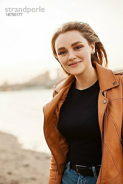 Glückliche junge Frau in modischer Lederjacke lächelt und schaut in die Kamera  während sie am Ufer eines Sees in einem windigen Abend steht
