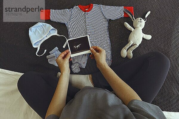 Cropped Schuss der schwangeren Frau Blick auf niedliche Babykleidung während auf dem Bett zu Hause ausruhen  weibliche werdendes Kind genießt Schwangerschaft  die Vorbereitung für die Elternschaft und Geburt. Adorable neugeborenen Kleidung