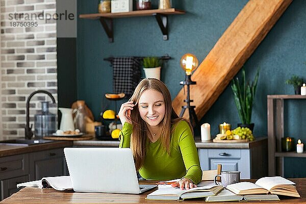 Junge  glückliche Freiberuflerin sitzt zu Hause am Tisch mit Laptop  macht sich Notizen  starrt nachdenklich in die Luft  denkt über neue Ideen nach  Bloggerin auf der Suche nach Inspiration. Freiberufliches Konzept