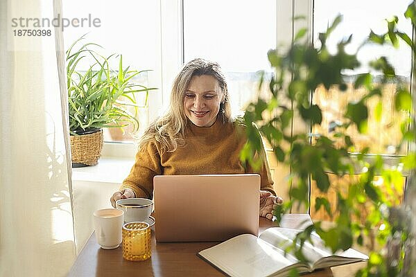 Fröhliche  glückliche Frau mittleren Alters  die zu Hause auf ihrem Laptop online chattet und einen Videoanruf über das Internet tätigt  überglückliche Frau in den 50ern  die auf den Computerbildschirm schaut und die Arme vor Glück ausbreitet  während sie am Tisch sitzt