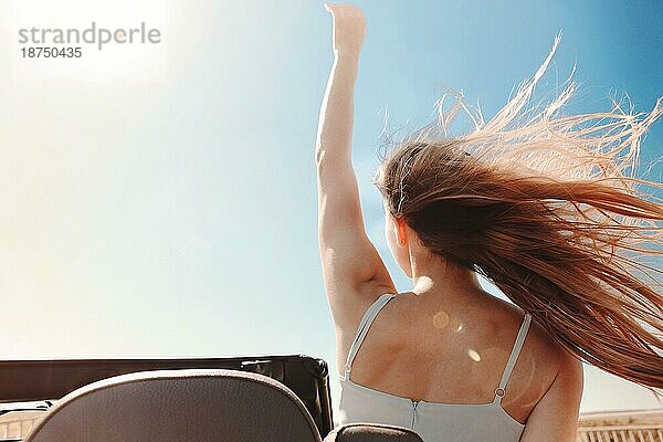 Rückenansicht einer fröhlichen jungen Frau in Sommerkleidung  die auf dem Beifahrersitz eines Cabriolets sitzt und während einer Autofahrt über die Schulter in die Kamera schaut