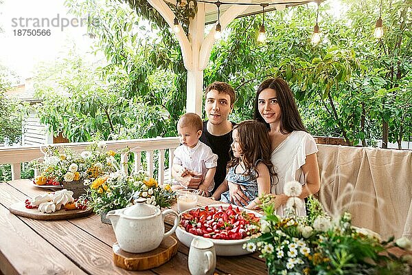 Liebevolle Familie mit Kindern am Tisch sitzend mit leckeren Desserts und Blumen beim Frühstück auf dem Land im Sommer