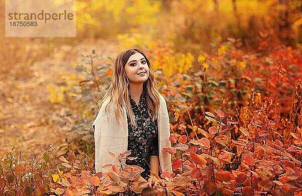 Schöne junge blonde Frau trägt Boho Stil langes Kleid beim Sitzen mit nachdenklichen Blick in hohem Gras im Herbst Wald in roten und gelben Farben dekoriert  Blick weg mit verträumten geheimnisvollen Lächeln