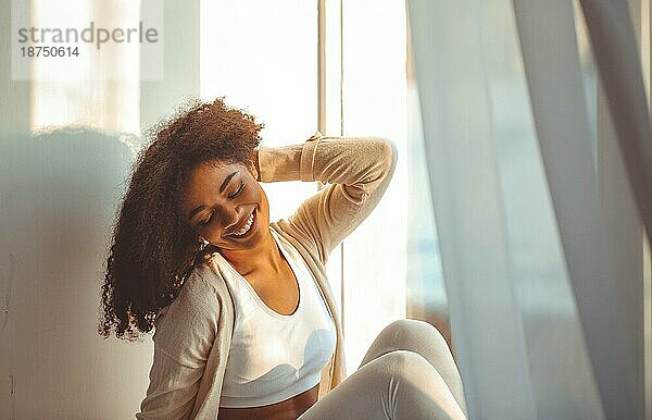Zufriedenes entspanntes afroamerikanisches Mädchen in Hauskleidung auf der Fensterbank sitzend vor dem Hintergrund der vom leichten Wind flatternden Vorhänge  glückliche Afro Frau zu Hause  die sanft mit geschlossenen Augen lächelt