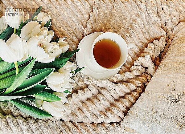 Strauß frischer weißer Tulpen und weiße Tasse Kaffee auf gemütlichem Bett mit beiger Plüschdecke und Kissen in hellem Schlafzimmer platziert
