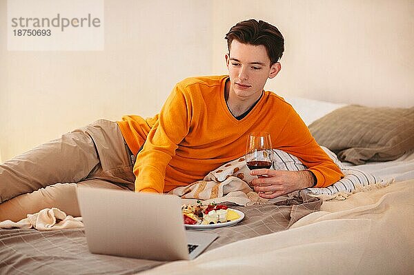 Entspannter junger Mann mit einem Glas Wein in der Hand  der auf einem bequemen Bett mit einem Teller Essen liegt und beim Essen Videos auf dem Laptop anschaut  während er sich zu Hause entspannt