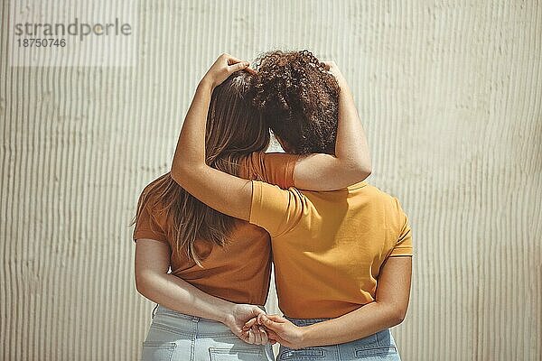 Starke weibliche Freundschaft. Rückansicht zwei Teenagermädchen beste Freunde halten Hände hinter dem Rücken und umarmen  während vor beige Wand im Freien stehen