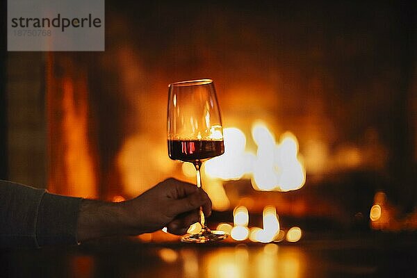 Mann mit Rotwein durch den Kamin. Close up von Mann hält und trinken Rotwein Glas von offenen Holzfeuer Platz entspannen und genießen ruhigen Moment zu Hause im Winterurlaub Urlaub  Weihnachten oder Freizeitkonzept