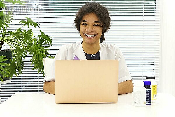 Junge glückliche afroamerikanische Ärztin  die am Arbeitsplatz sitzt und in die Kamera schaut und lächelt  während sie an einem Laptop in einer modernen Klinik arbeitet  zufriedener ethnischer Arzt  der am Tisch sitzt und die Arbeit im Krankenhaus genießt