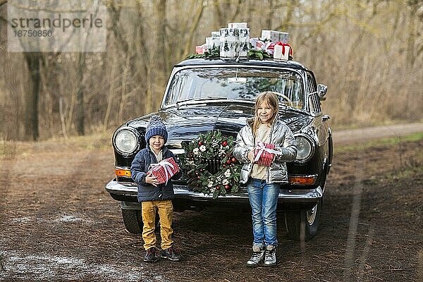 Charmante stilvolle Mädchen und Jungen geben Geschenke zu einander stehen in der Nähe von Retro schwarzes Auto mit Boxen auf dem Dach im Winter Park