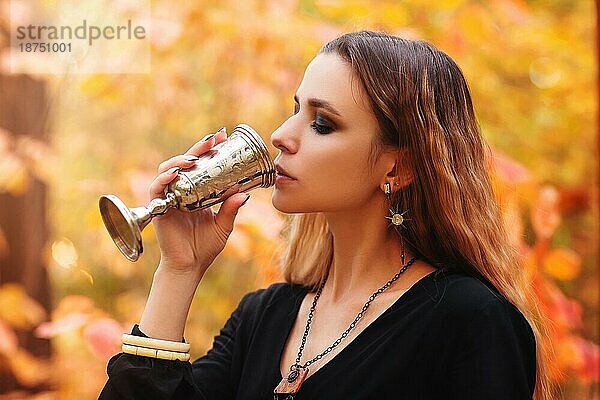 Junge fröhliche Frau in schwarzem Kleid hält Vintage Metall Glas in den Händen  während im Herbst Park mit bunten Blättern auf dem Hintergrund stehen  trinken Wein auf Picknick im Wald