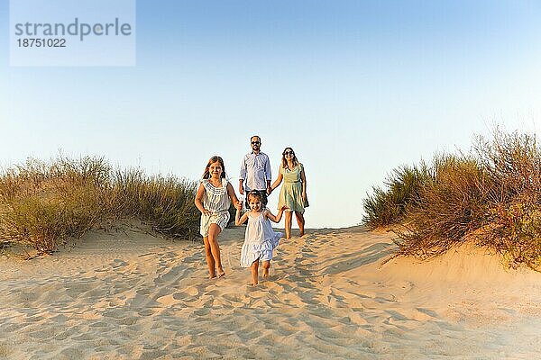 Glückliche vierköpfige Familie  junge Eltern und kleine Kinder  die den Sommerurlaub in der Natur genießen  am Sandstrand spazieren gehen  sich an den Händen halten  die Schwestern laufen voraus  während die Eltern hinterherlaufen