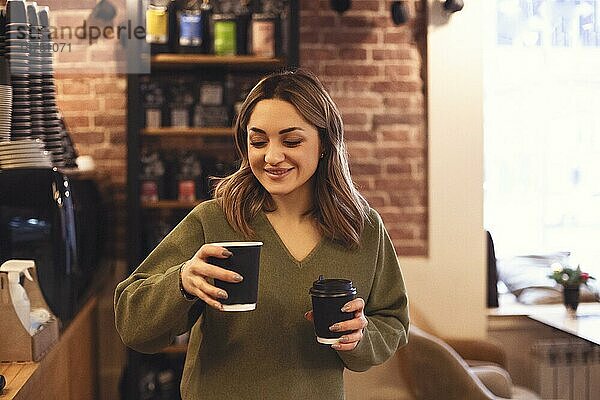 Innenaufnahme einer jungen lächelnden Frau  die zwei Tassen Kaffee zum Mitnehmen hält  während sie in einem Café steht  selektiver Fokus auf eine Frau  die heiße Getränke während eines morgendlichen Treffens mit einem Freund bringt