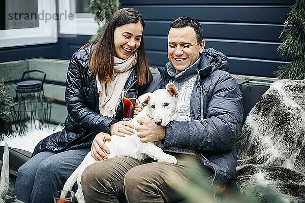 Junges Paar umarmt ihren weißen süßen Haushund. Mann und Frau auf Neujahrsmesse  trinken Glühwein  glücklich und lächelnd