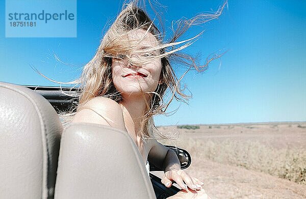 Fröhliche junge Frau in Sommerkleidung  die auf dem Beifahrersitz eines Cabrios sitzt und während einer Autofahrt über die Schulter in die Kamera schaut
