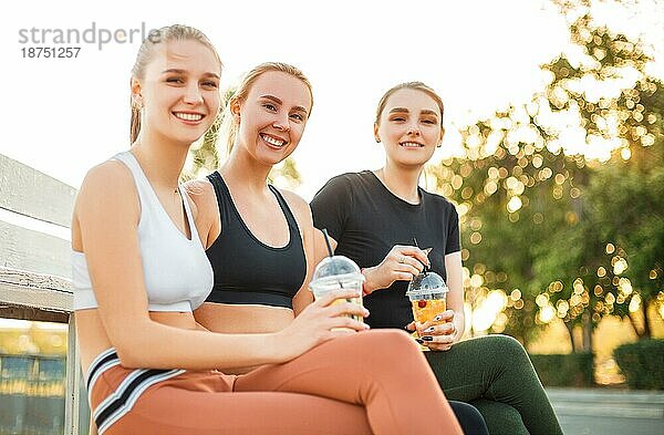 Junge Frau in Sportkleidung lächelt und schaut weg  während sie ein frisches  gesundes Getränk genießt und auf einer Bank in der Nähe von Freundinnen sitzt