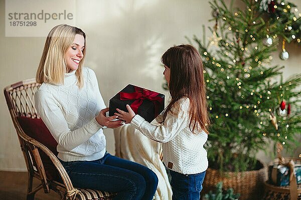 Nettes Mädchen Tochter geben Weihnachtsgeschenk ihre junge lächelnde Mutter  in einem Raum mit geschmückten Baum sitzen  glückliche Familie Mutter und Kind tragen weiße gestrickte Pullover feiern Neujahr zu Hause