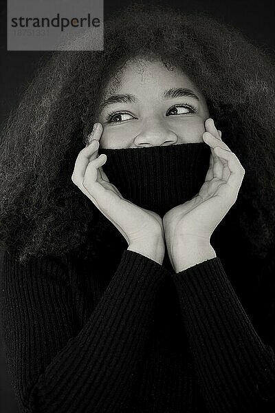 Zufriedene afroamerikanische lockige Frau  die die Augen schließt und das Gesicht mit einem warmen schwarzen Pulloverkragen bedeckt  vordunklem Studiohintergrund  lächelnde Frau  die sich in einem Rollkragenpullover aufwärmt. Vertikales Foto. Schwarz und weiß